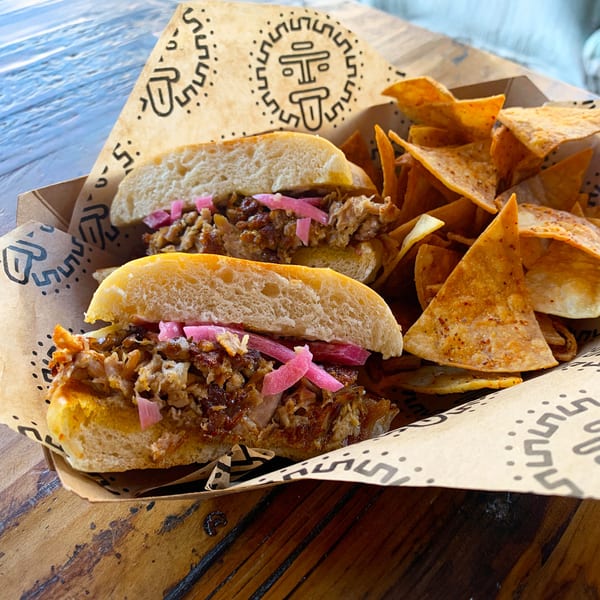 Two Taqueria Picoso carnitas sandwiches with pickled onions and a side of tortilla chips.
