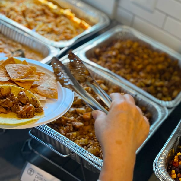 A hand uses tongs to serve Mexican food from a buffet into a taco with tortilla chips.