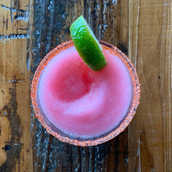 Overhead view of a pink frozen margarita with a lime wedge and a chili-salt rim on a wooden table.