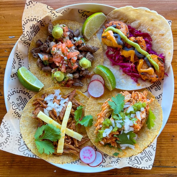 A white plate holds four colorful Taqueria Picoso tacos with various fillings, garnishes, and lime wedges.