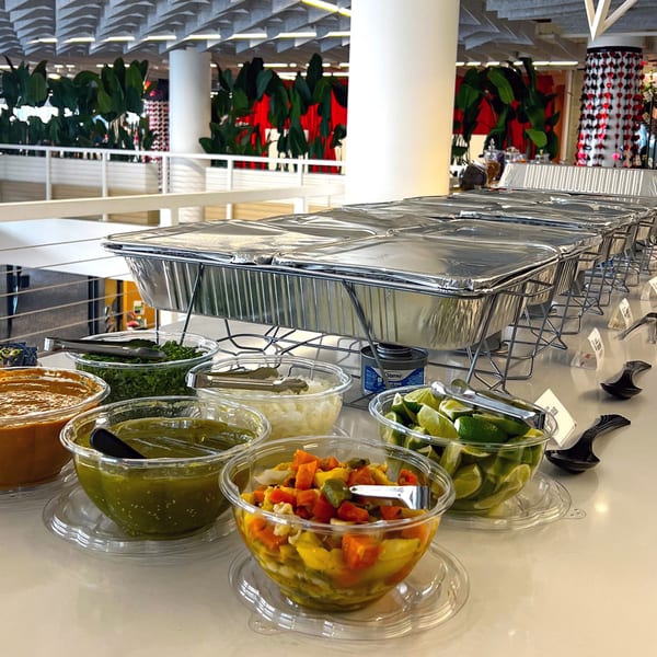 A buffet line with foil-covered food warmers and bowls of Mexican toppings like salsa, limes, and onions.