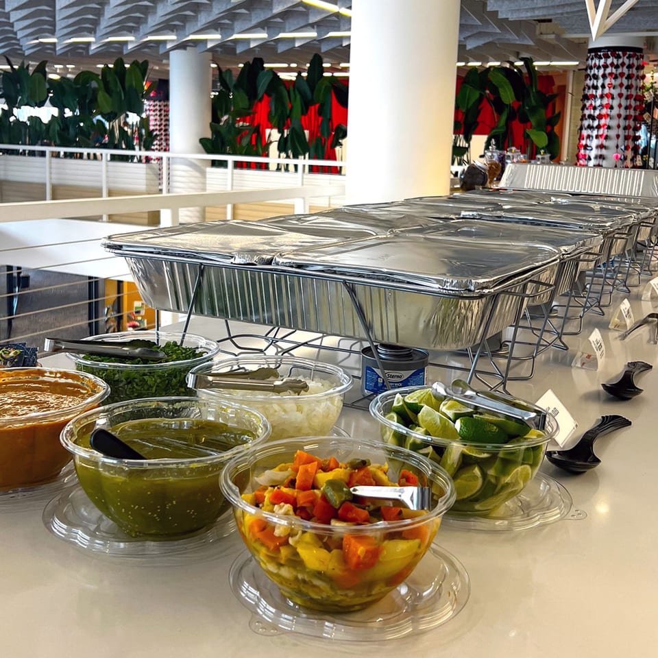 A buffet line with foil-covered food warmers and bowls of Mexican toppings like salsa, limes, and onions.