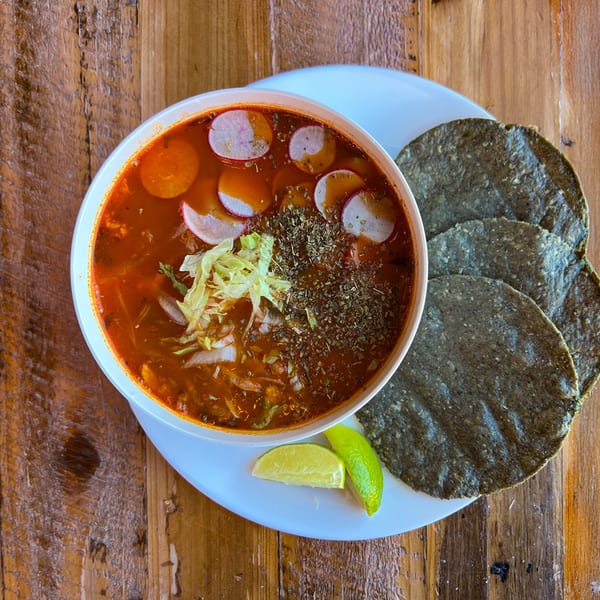 A bowl of Taqueria Picoso's red pozole with radishes, lettuce, and herbs, served with blue corn tortillas and lime wedges.