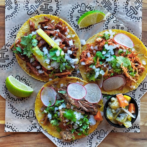 Three Taqueria Picoso tacos with various fillings, cilantro, onions, radishes, and lime wedges.